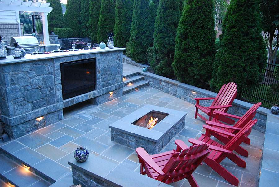 A stone pavers patio with a fireplace, outdoor kitchen, and four red lawn chairs.