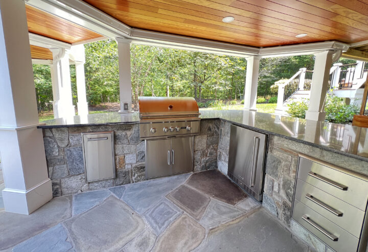 An outdoor kitchen with the marble-top counter formed into an L-shape underneath a wooden pavilion