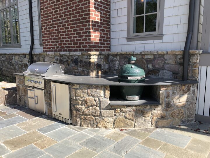 An outdoor kitchen with stone siding equipped with a grill, fridge, and green egg smoker