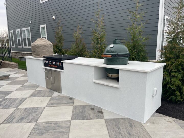 A white outdoor kitchen counter with a stovetop, green egg smoker, and a pizza oven
