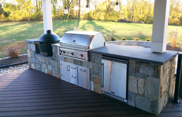 An outdoor kitchen with a green egg smoker, grill, and fridge positioned at porch level.